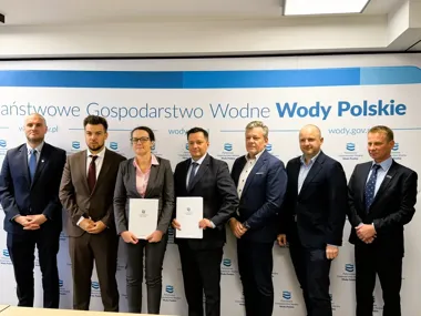 Grupa osób w garniturach pozuje do zdjęcia przed ścianką Państwowego Gospodarstwa Wodnego Wody Polskie, dwie osoby trzymają dokumenty.