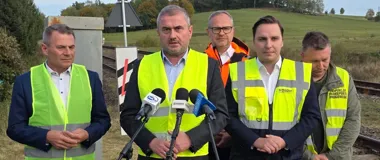 Mężczyźni w kamizelkach odblaskowych stoją przy torach kolejowych podczas konferencji prasowej na otwartym terenie.