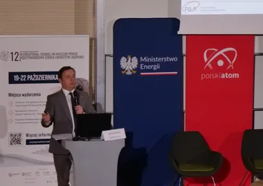 Mężczyzna w garniturze przemawia z pulpitu podczas konferencji poświęconej energetyce jądrowej, w tle banery Ministerstwa Energii i Polskiego Atomu.