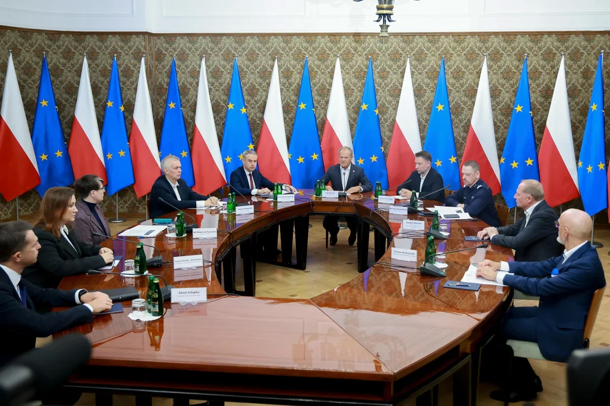 Spotkanie przy owalnym stole w reprezentacyjnej sali z flagami Polski i Unii Europejskiej w tle.