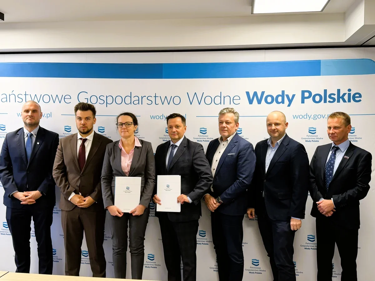 Grupa osób w garniturach pozuje do zdjęcia przed ścianką Państwowego Gospodarstwa Wodnego Wody Polskie, dwie osoby trzymają dokumenty.